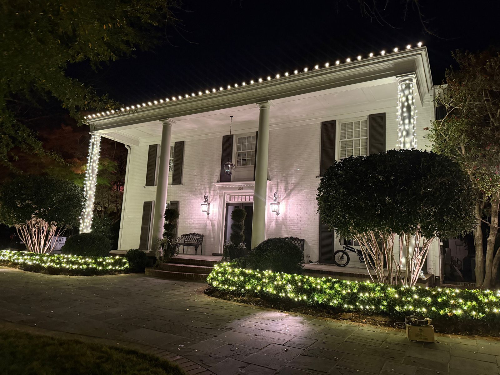 Custom-cut warm white Christmas lights running in a perfectly even line along the roofline of a white colonial home in Raleigh, NC, with wrapped columns and netted shrubs