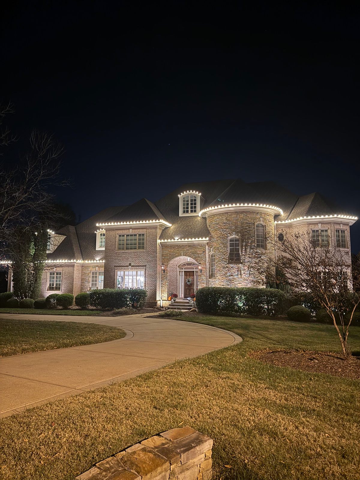 Brick and stone home with warm white Christmas lights tracing every gable, dormer, and the round stone turret